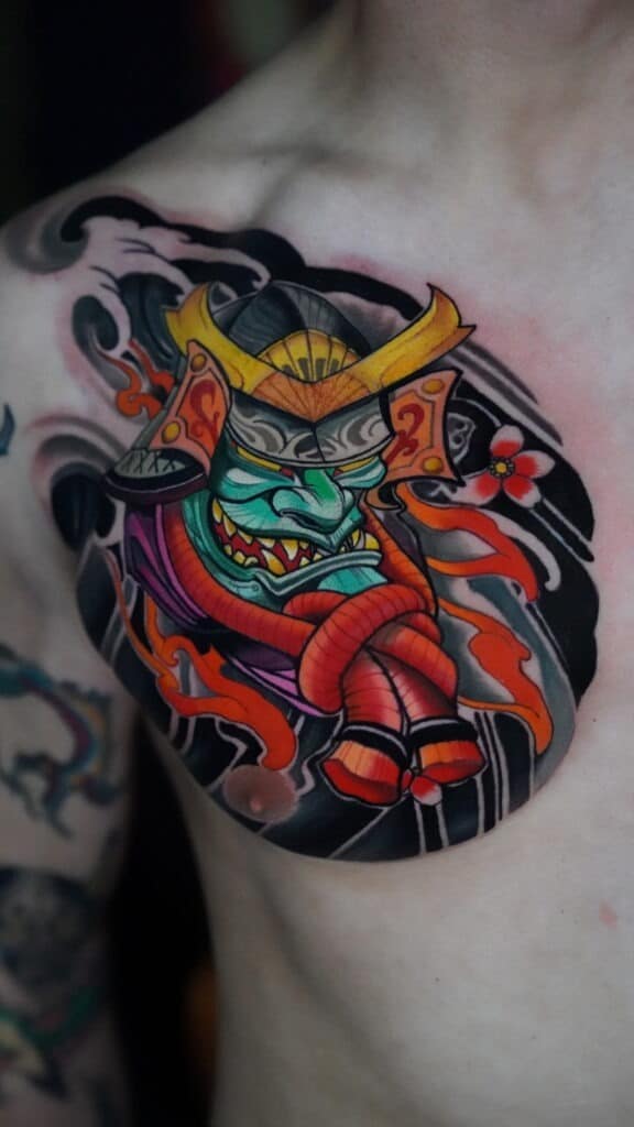 Colorful tattoo of a snarling samurai mask with a horned helmet, red rope, flames, and a cherry blossom