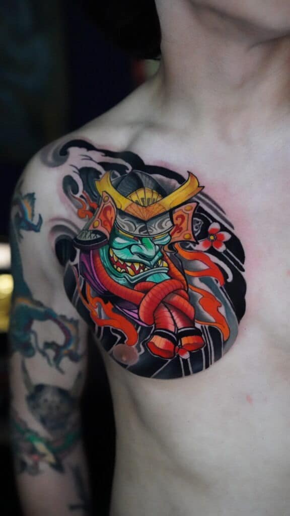 Colorful chest tattoo of a samurai mask with horns, red rope, black waves, and a small flower