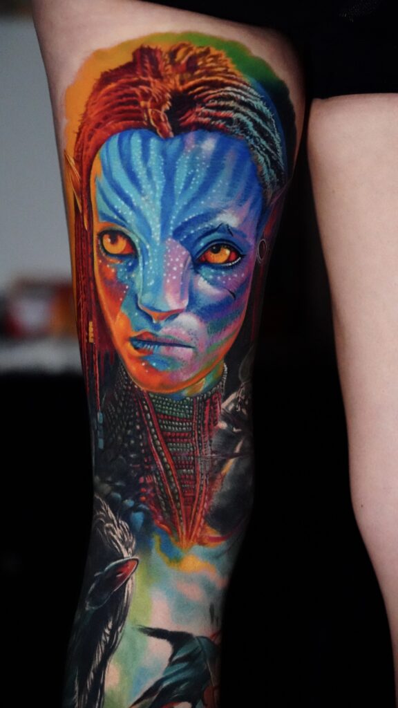 Colorful forearm tattoo of a blue Na'vi face with yellow eyes and braided hair