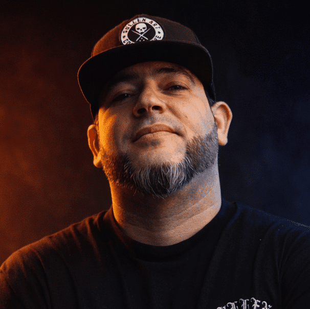 Portrait of a bearded man wearing a black cap and black shirt, lit with orange and blue light