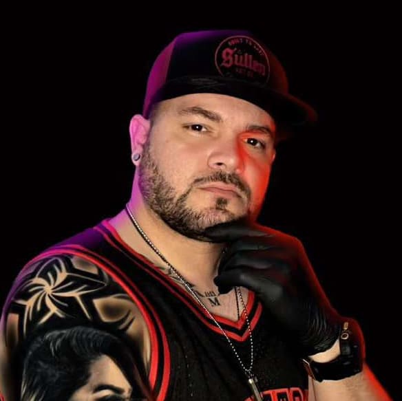Man wearing a black cap and glove, posing with hand on chin, showing a tattooed shoulder